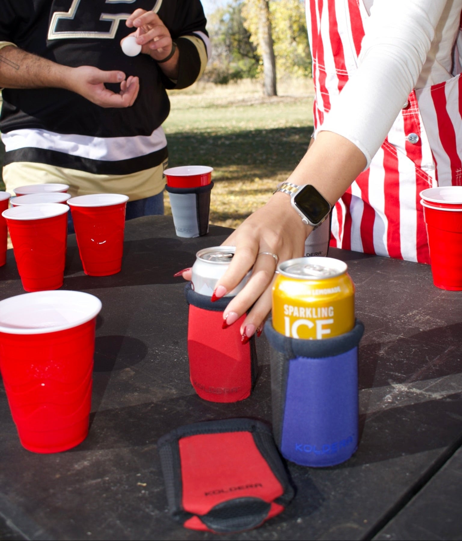 Two people at a table with red cups and colorful can coolers outdoors.